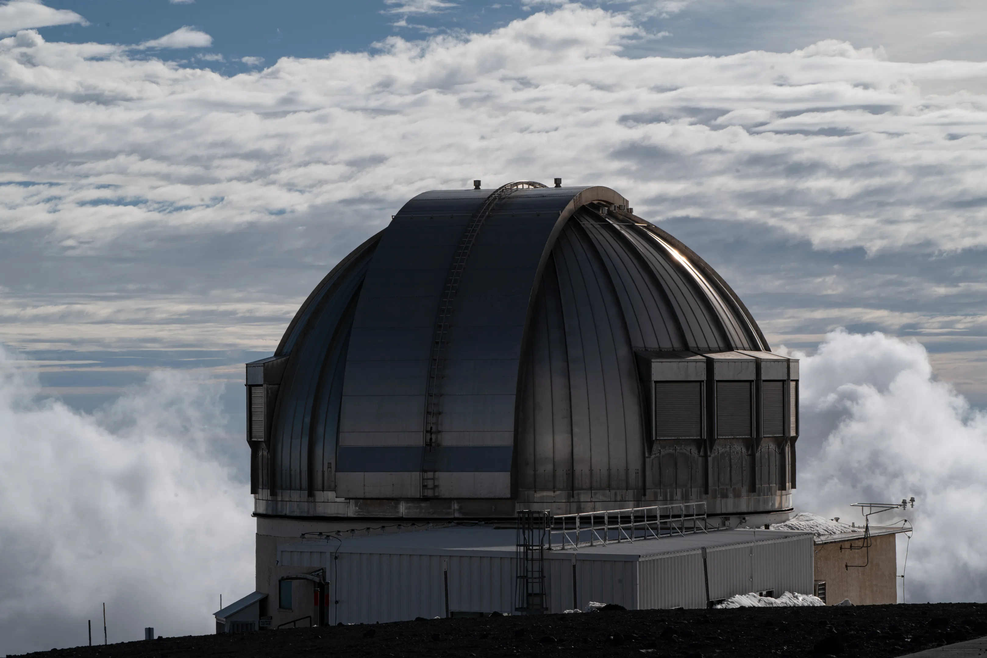 An astronomical observatory dome on the summit of Mauna Kea, above a sea of clouds — a metal dome on a field of dark lava with patches of snow.